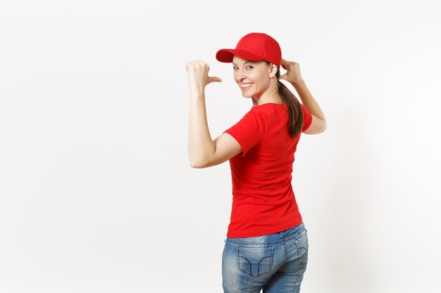 Photo delivery woman in red uniform isolated on white background. professional pretty female in cap, t-shirt, jeans working as courier or dealer, pointing fingers on copy space for advertisement. back view.