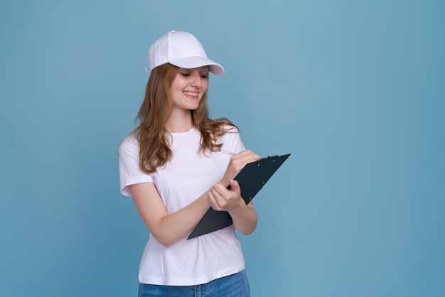 Delivery woman writing document isolated on blue background in white tshirt and cap Caucasian cheerful girl moonlights as a promoter and consultant