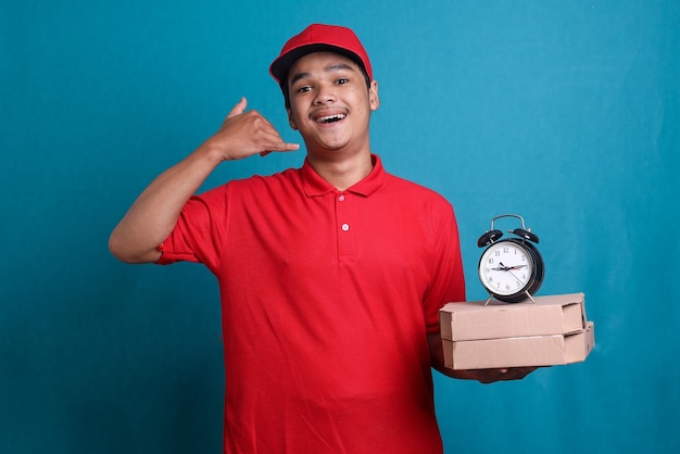 Delivery worker in red uniform is holding boxes of pizza and a clock while doing call gesture. On ti