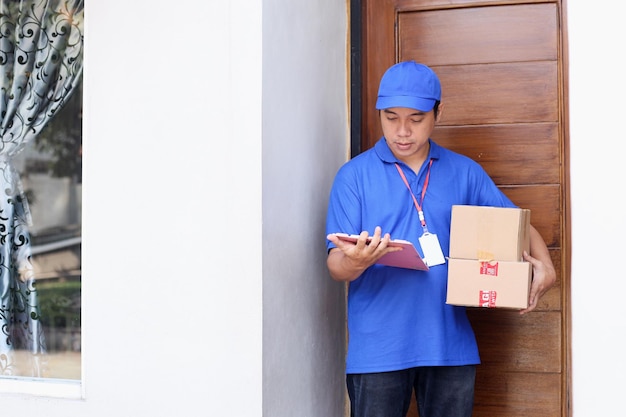 Deliveryman in blue uniform holding box and clipboard, checking shipment status in front of the door