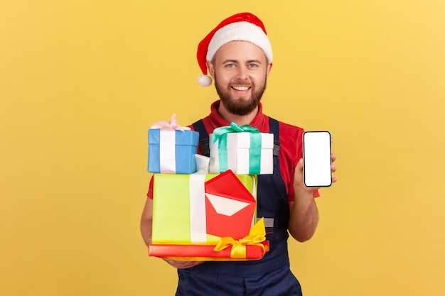 Deliveryman in red santa claus hat holding holidays gifts and showing smartphone with blank display