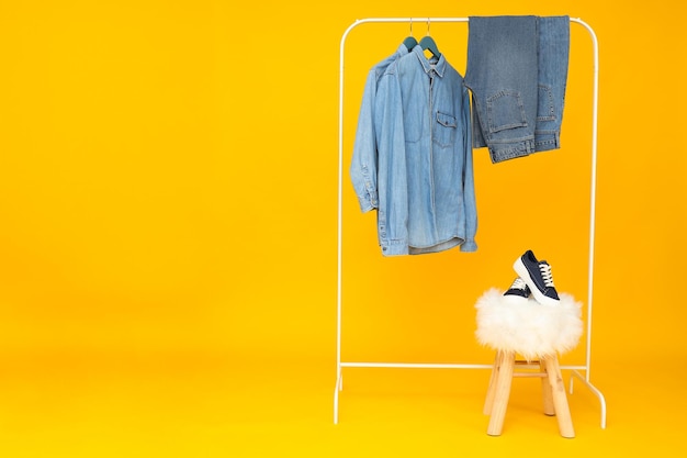 Denim pants and shirts on hangers shoes on stool on yellow background space for text