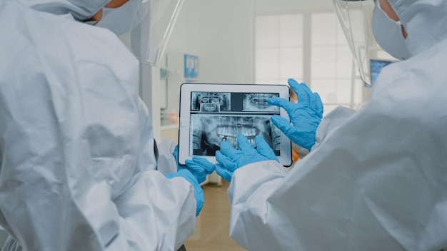 Dental doctors examining x ray of teeth on tablet screen at stomatology clinic. Professional dentists with hazmat suits looking at digital device used for dentition model implant