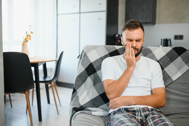 Photo dental health problems concept man suffering from strong toothache upset male patient with inflamed nerve tooth abscess or teeth sensitivity sitting on sofa and touching cheek with grimace of pain