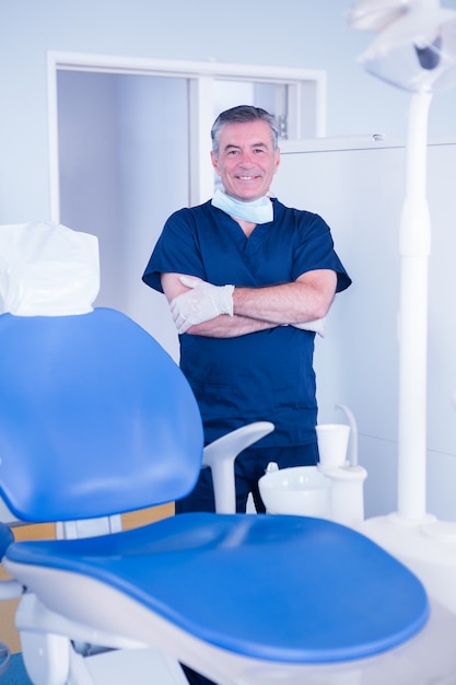 Dentist in blue scrubs smiling at camera beside chair