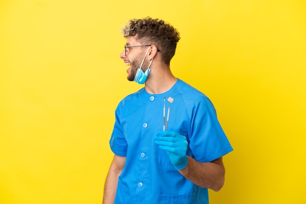 Dentist caucasian man holding tools isolated on yellow background laughing in lateral position