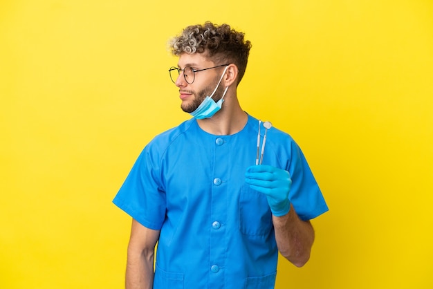 Dentist caucasian man holding tools isolated on yellow background looking to the side