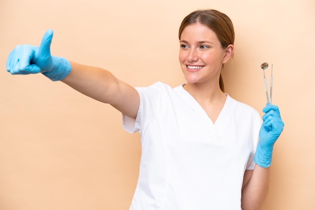 Dentist caucasian woman holding tools isolated on beige background giving a thumbs up gesture