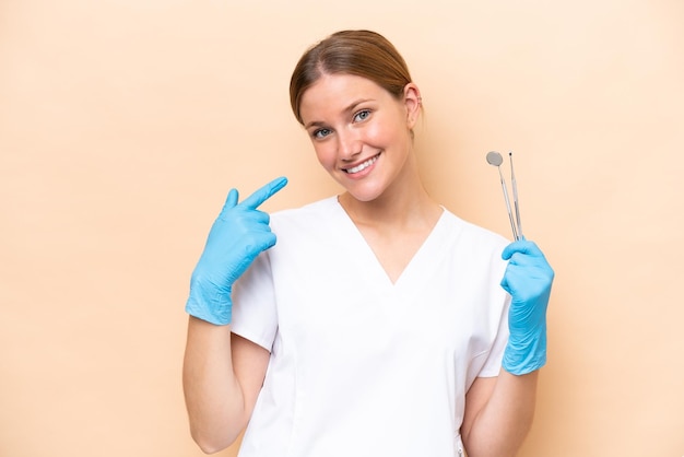 Dentist caucasian woman holding tools isolated on beige background giving a thumbs up gesture