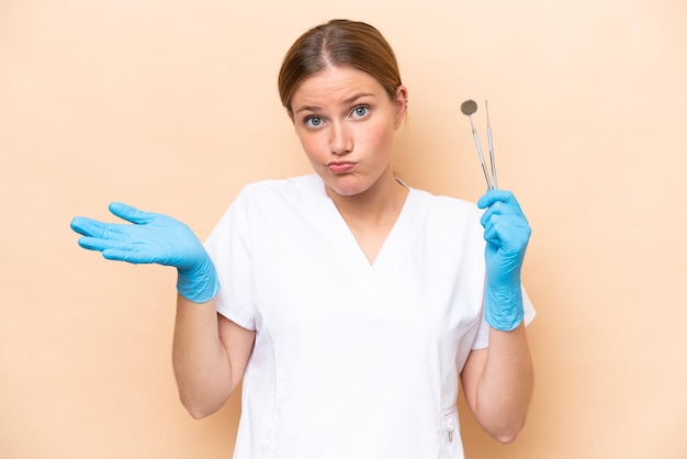 Dentist caucasian woman holding tools isolated on beige background having doubts while raising hands