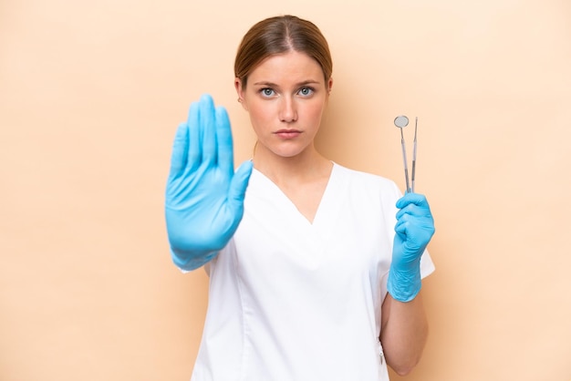 Dentist caucasian woman holding tools isolated on beige background making stop gesture