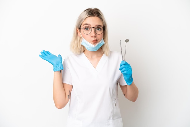 Dentist caucasian woman holding tools isolated on white background having doubts while raising hands