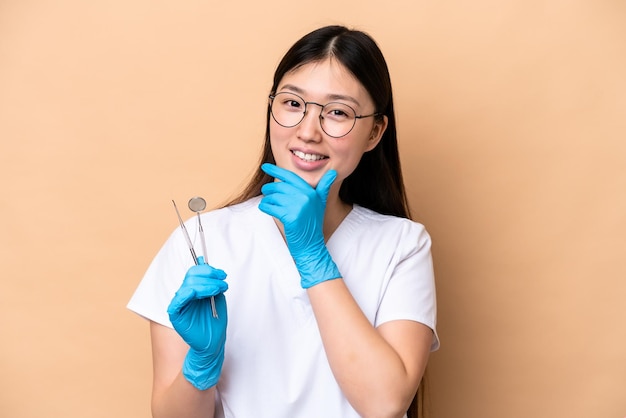 Dentist Chinese woman holding tools isolated on beige background happy and smiling