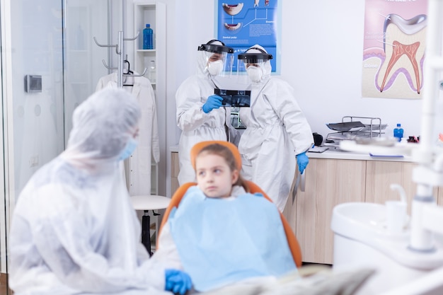 Dentist doctor and assistant holding dental radiography of little kid patient in the course of coronavirus. Stomatologist during covid19 wearing ppe suit doing teeth procedure of child sitting on chai