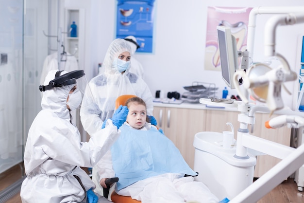 Dentist doctor in the course little kid consultation in dentistery office. Stomatolog in protectie suit for coroanvirus as safety precaution holding child teeth x-ray during consultation.