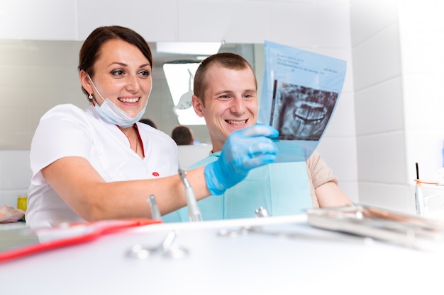 Dentist finishing successful teeth restoration, black haired doctor, in green mask, giving to happy patient mirror, beautiful long-haired woman looking at pristine white teeth with great pleasure