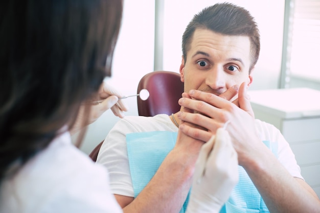 A dentist is trying to give a treatment to a man but she canÃ¢ÂÂt because he is extremely scared and shows it with his hands over his mouth and a look on the instruments