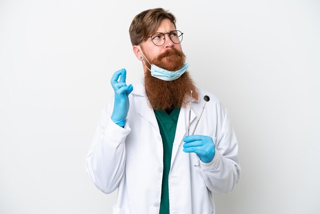 Dentist reddish man holding tools isolated on white background with fingers crossing and wishing the best