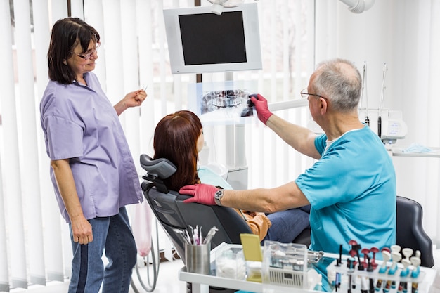 Dentist team, senior man dentist and his female assistant, in dental office talking with female patient and preparing for treatment, while examining of x-ray image