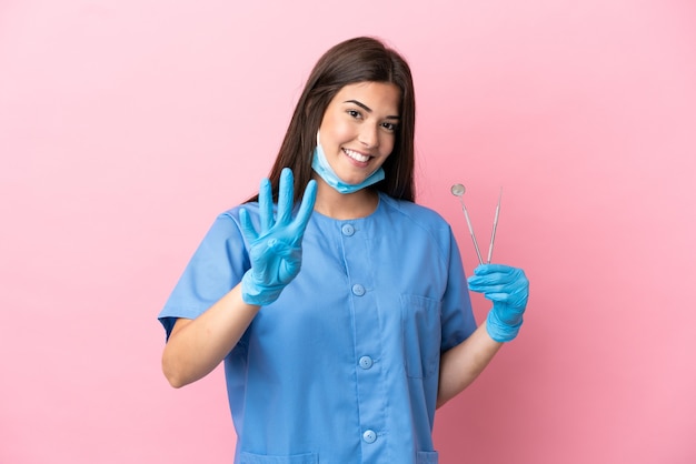 Dentist woman holding tools isolated on pink background happy and counting four with fingers