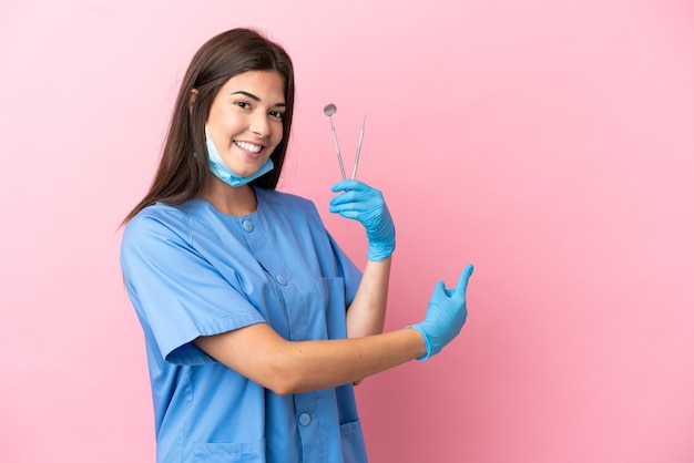 Dentist woman holding tools isolated on pink background pointing back