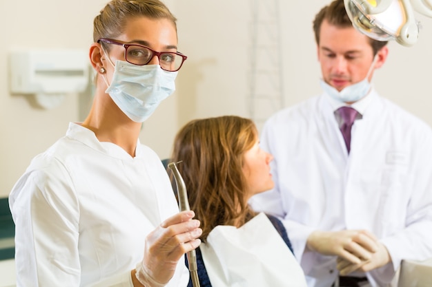 Dentistshis surgery holds a drill and looking at the viewer, in the  her colleague is giving a female patient a treatment