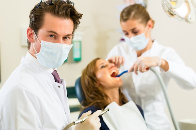 Dentistshis surgery looking at the viewer, in the  his assistant is giving a female patient a treatment