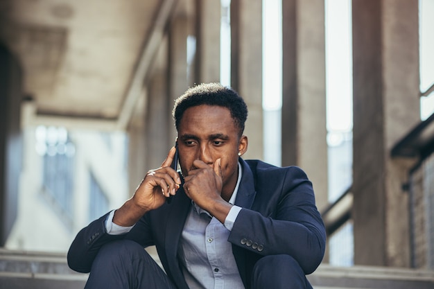 Depressed african american businessman telling sad news talking on cell phone sitting on office stairs in business suit