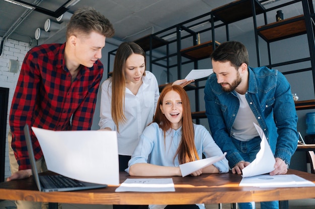 Depressed angry young business woman under stress throwing documents at desk looking at camera multiethnic colleagues standing near her