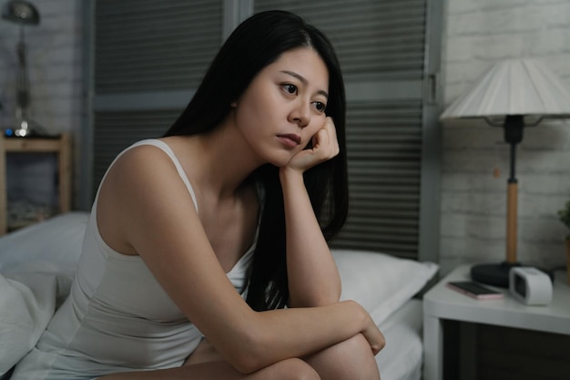 Depressed asian korean woman awake in night. young female wear tank top feeling exhausted and suffering from insomnia while sitting in white cozy bed. sad lady looks pensive think problem in bedroom