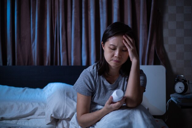 Depressed Asian woman can not sleep on the bed. Worry girl sitting and holding drugs for check detail before using in the bedroom.Sickness pain after headache and ill healthy.