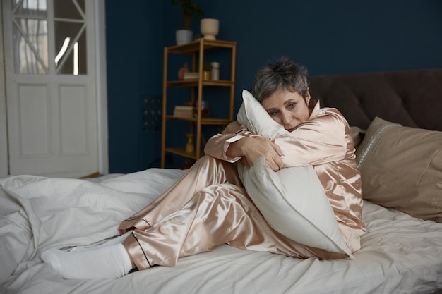 Depressed elderly woman hugging pillow sitting on bed