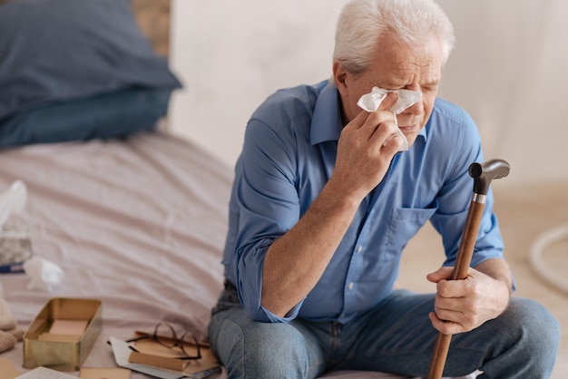 Depressed gloomy elderly man crying and wiping away his tears while holding a paper tissue