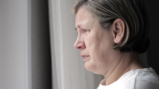 Depressed mature woman cries standing near window in bedroom