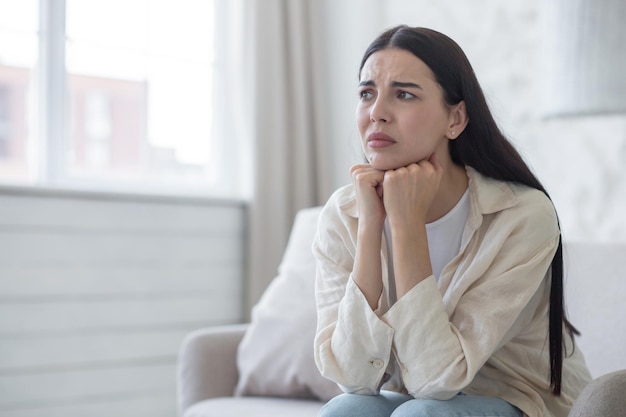 Depressed woman near window sitting on sofa at home disappointed and sad brunette grieving divorce