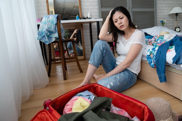 depressed woman sitting on wooden floor in bedroom packing after breaking up with boyfriend. frowning asian unhappy girl next luggage going to travel trip alone with breaking heart relax enjoy.