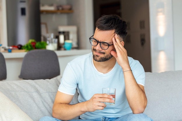 Depressed young man having headache while drinking water on sofa at home