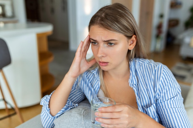 Depressed young woman having headache while drinking water on sofa at home