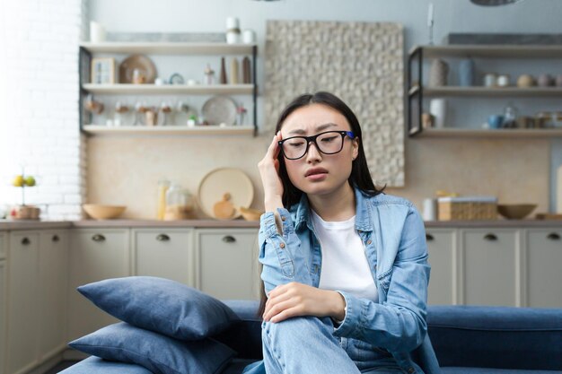 Depression tired and pensive young asian woman sitting on sofa at home holding hand to head looking