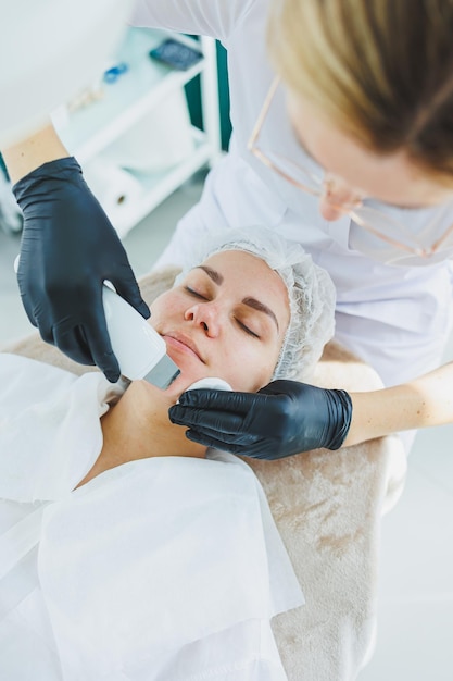 A dermatologist or cosmetologist in a clinic works with female patients and makes facial skin procedures on a young womans face Ultrasonic cleaning of the face in the cosmetologists office