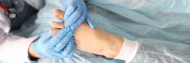 Dermatologist is holding foot of woman with damaged thumb in his hands in rubber gloves in clinic