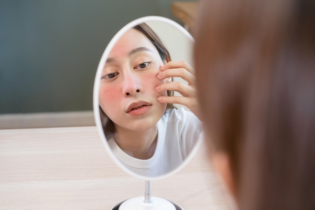 Dermatology puberty asian young woman girl looking into mirror allergy presenting an allergic reaction from cosmetic red spot or rash on face Beauty care from skin problem by medical treatment