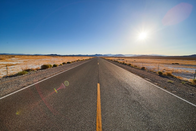 Desert paved road leading into endless desert with sun and blue sky