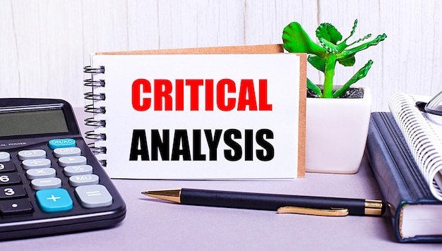 On the desktop are a calculator, diaries, a potted plant, a pen and a notebook with the text CRITICAL ANALYSIS. Business concept. Workplace close up