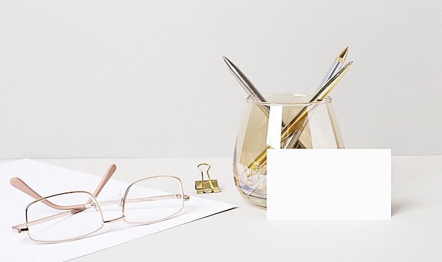 On the desktop are gold glasses, paper clips, pens in a glass beaker, and a white blank card with space to insert text. Business template. Home Office. Close-up of the workplace.