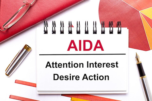 On the desktop is a white notebook with the text AIDA Attention Interest Desire Action a pen burgundy and red tables and goldframed glasses Business concept