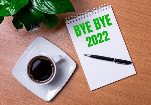 On the desktop there is a green plant a white cup of coffee a pen and an open notepad with the text BYE BYE 2022 Business concept Closeup of the workplace