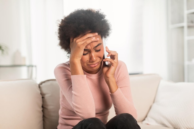 Despaired sad depressed young mixed race lady sitting on sofa calling by smartphone presses hand to head