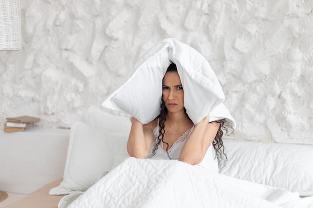 Despaired sad young caucasian brunette female in pajamas covers her ears with pillow sits on bed