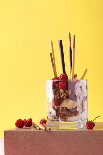 Dessert in glass with chocolate granola, banana and raspberry, decorated with sticks Pocky on pink and yellow background, Vertical format, Space for text